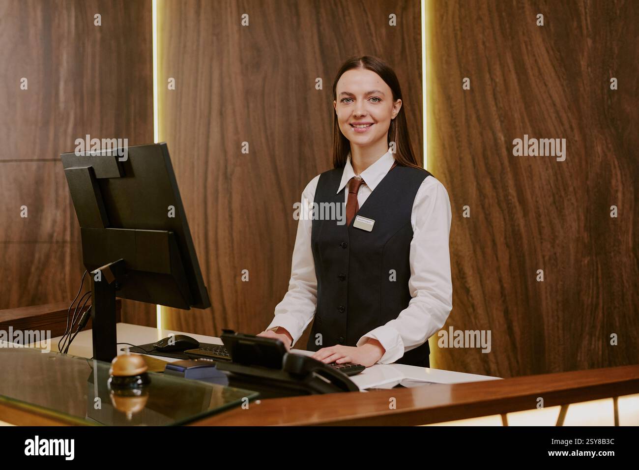 Medium close up of adorable smiling receptionist standing near table with computer while working at hotel Stock Photo