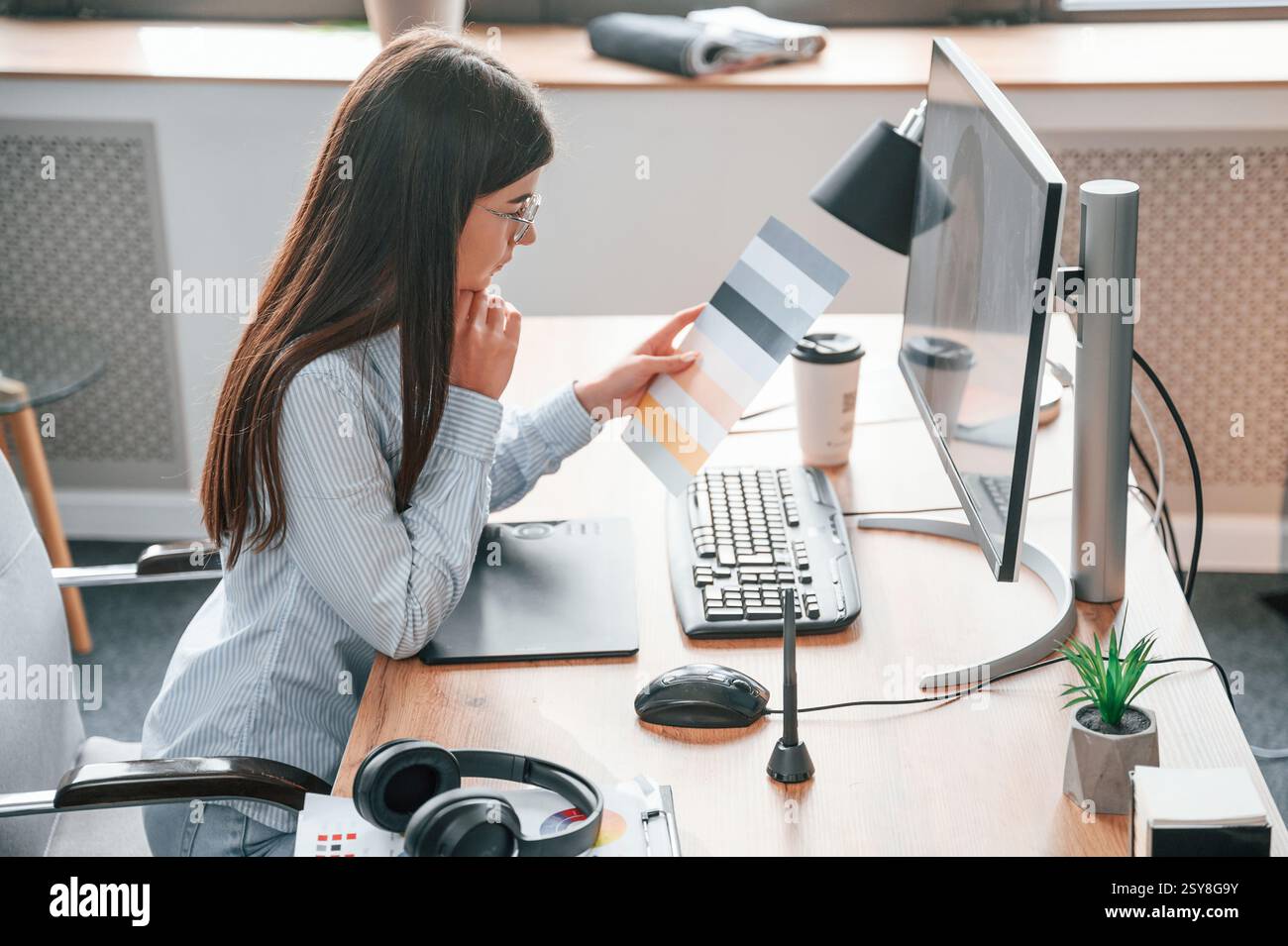 SIde view. Sitting by computer. Young female designer is in the modern office. Stock Photo