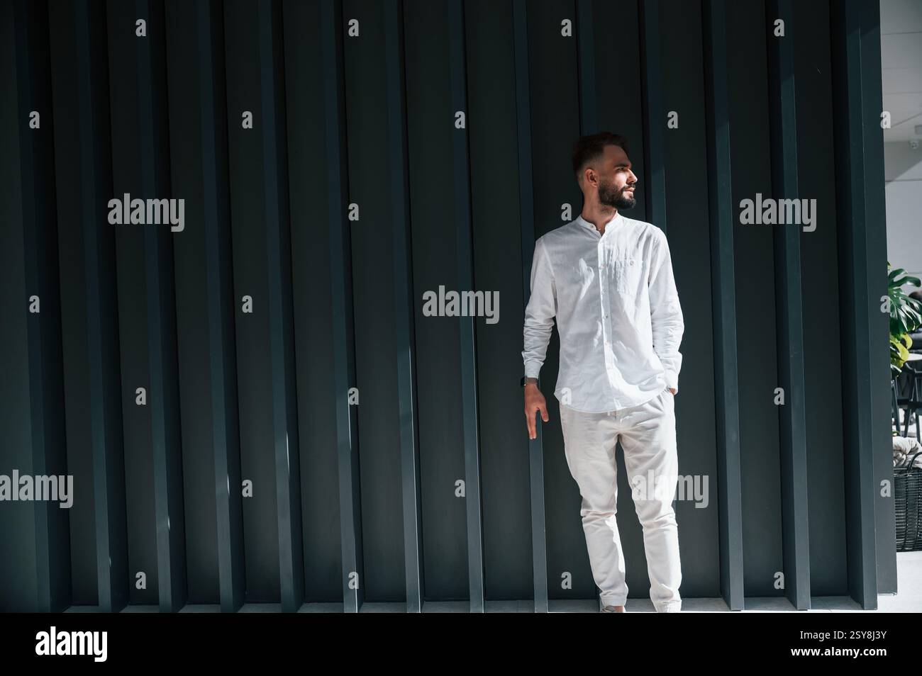 Young handsome man with beard and in white shirt is standing against dark green background. Stock Photo