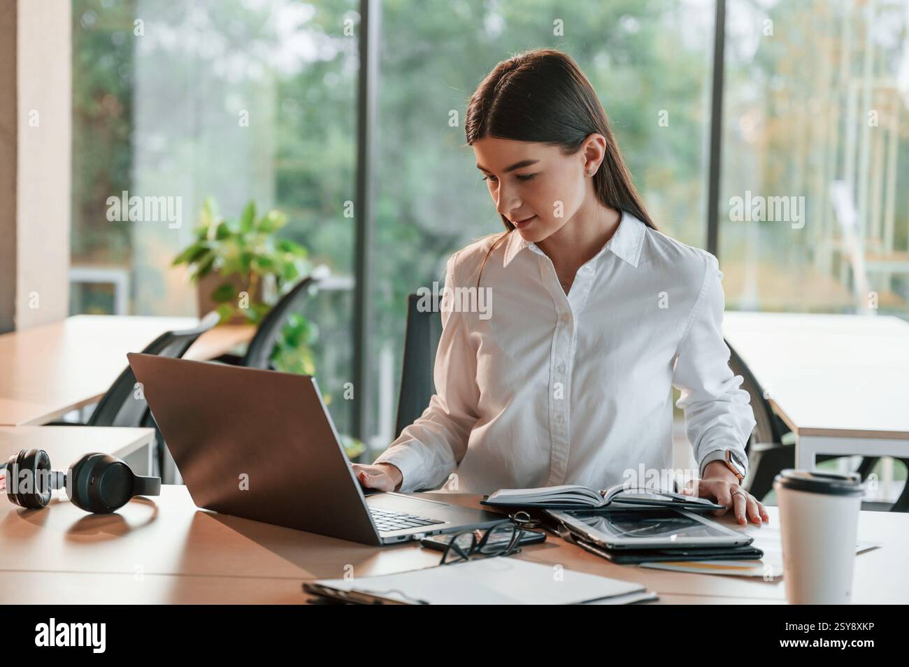 With laptop. Beautiful young woman in white clothes is working in office. Stock Photo