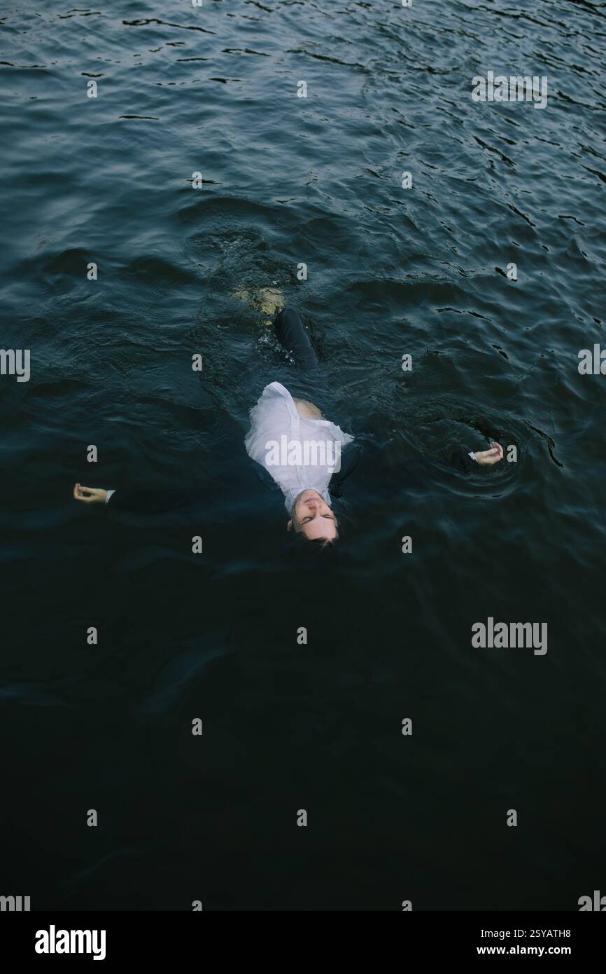 A man dressed in a suit floats on dark water with arms outstretched ...