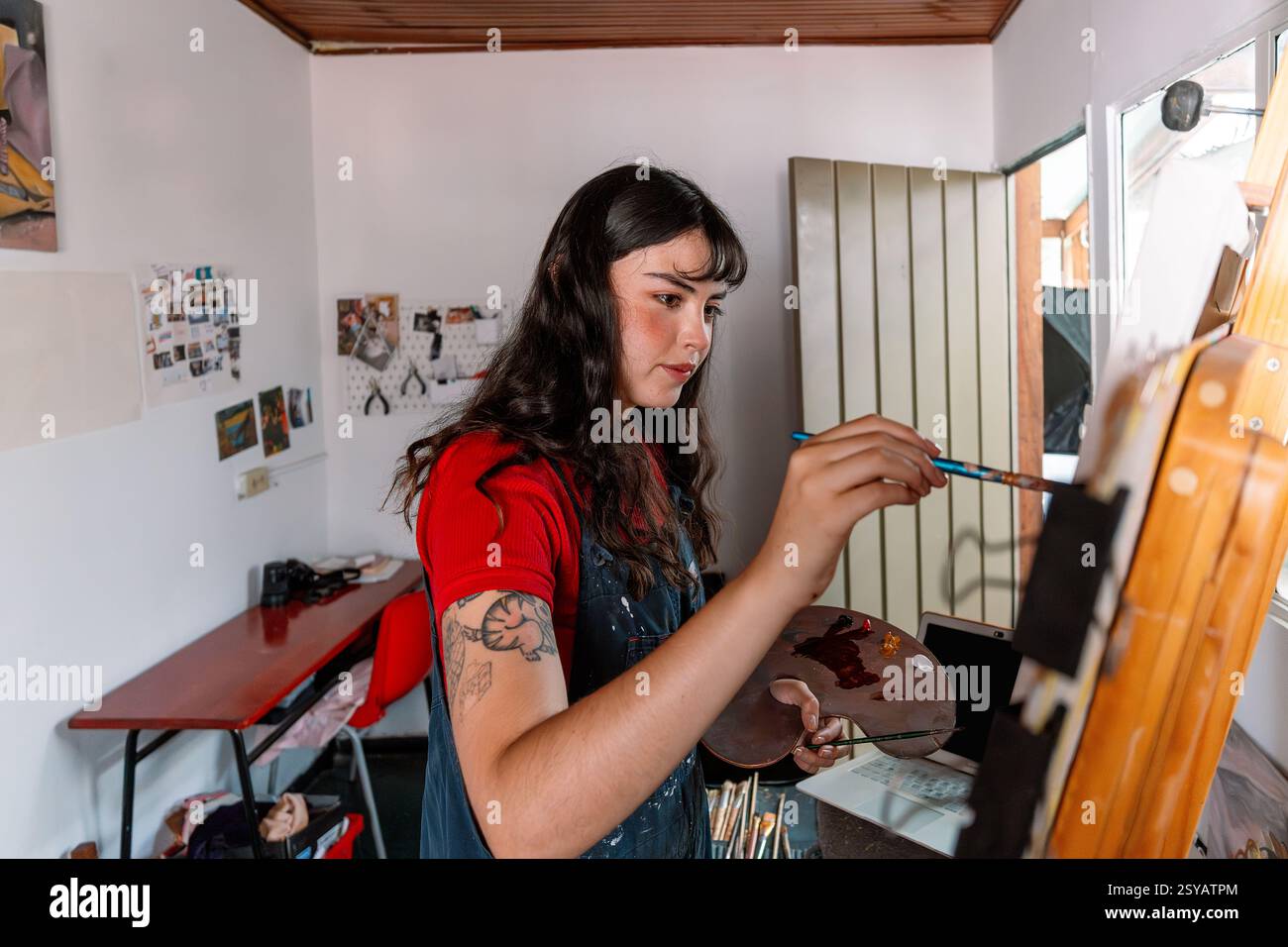 A young woman artist is focused on painting a canvas in her home studio ...