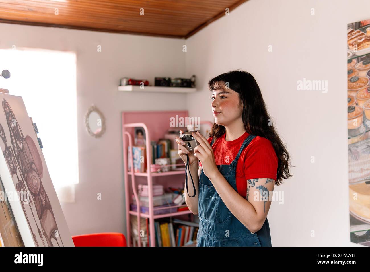 A young woman in a red shirt and paint-splattered overalls holds a vintage camera while standing in her creative studio. Shelves with books, cameras, Stock Photo