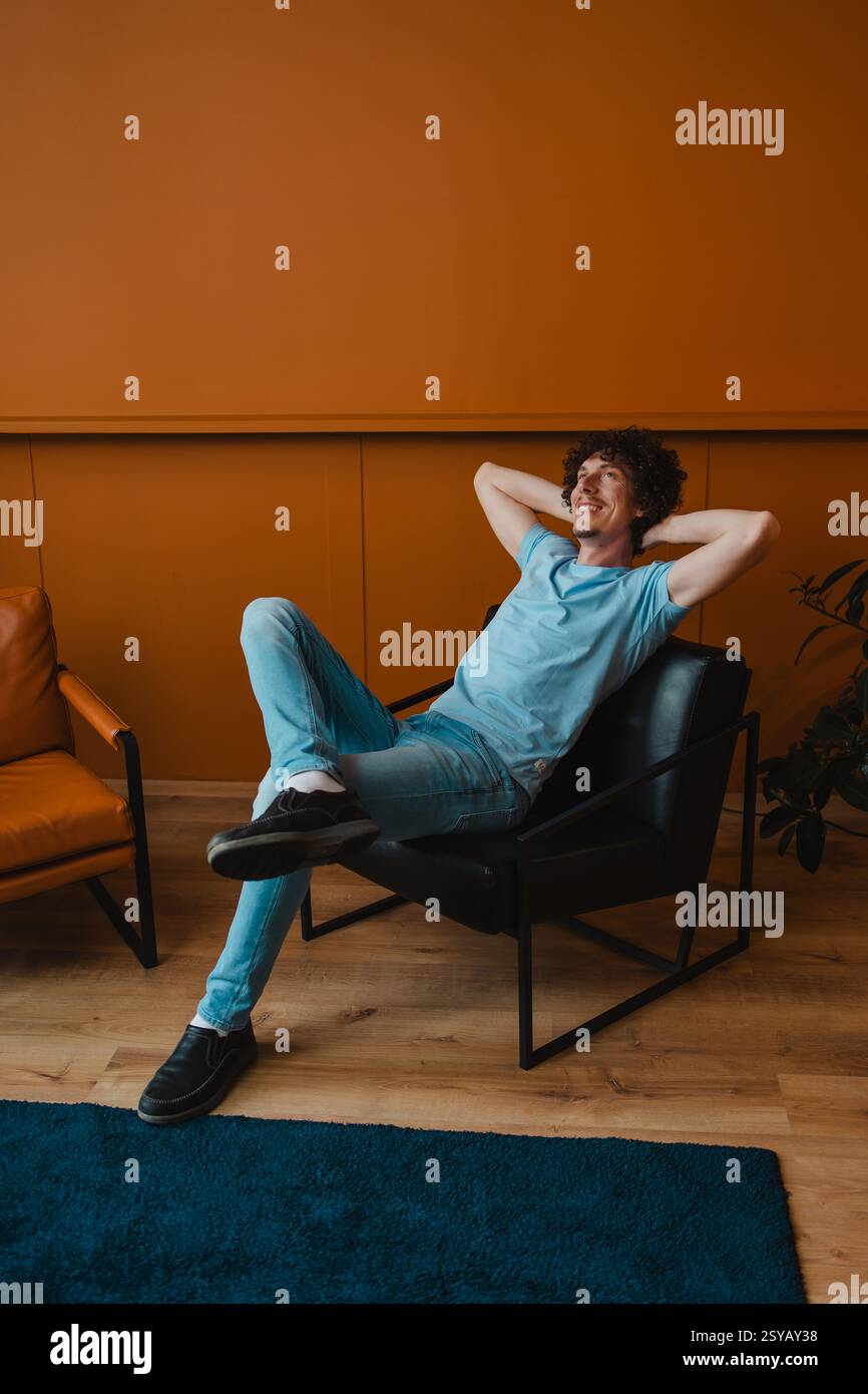 A young man with curly hair relaxes comfortably in a modern chair. His casual attire complements the stylish interior setting, creating a laid-back at Stock Photo