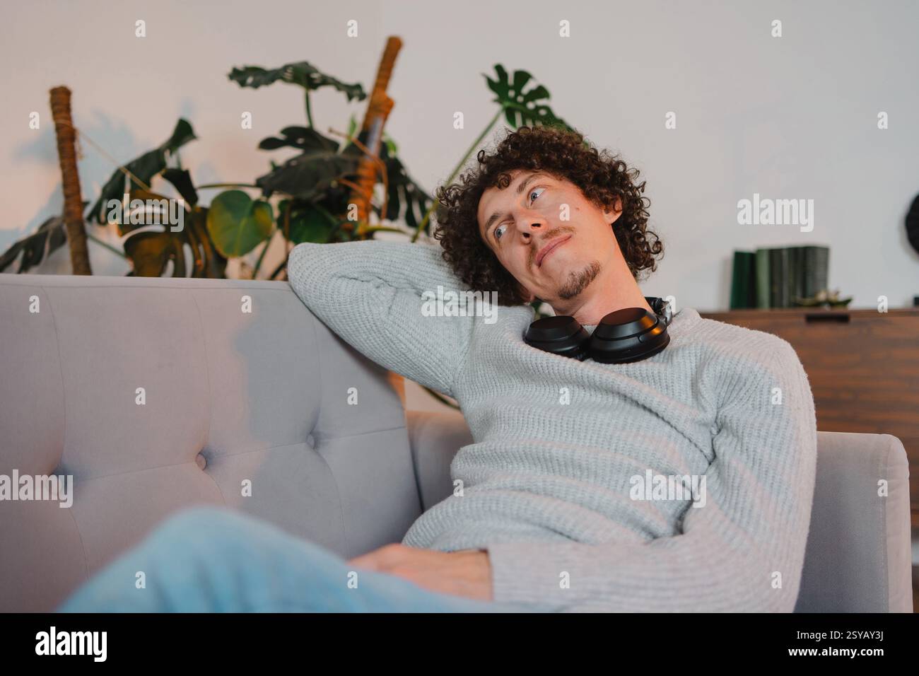 A curly-haired young man with casual attire, relaxing on a sofa with headphones around his neck. The serene setting features indoor plants in the back Stock Photo