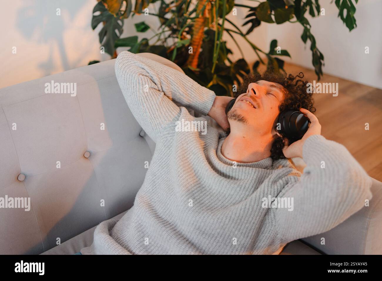 A young man with curly hair relaxes on a sofa, listening to music with headphones. Sunlight streams through a window, plants in the background, creati Stock Photo