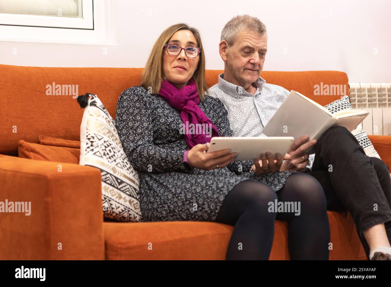 A couple with intellectual disabilities enjoys a evening indoors of a supervised home. The woman uses a tablet while the man reads a book. They sit on Stock Photo