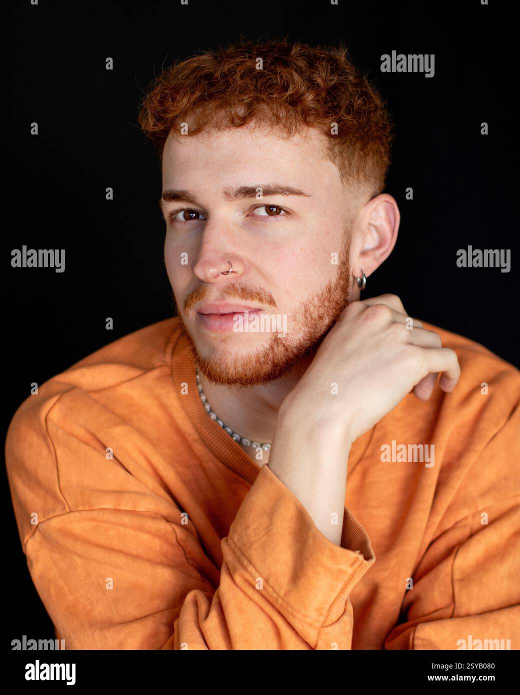 A young man with red hair and beard wearing an orange sweater poses ...