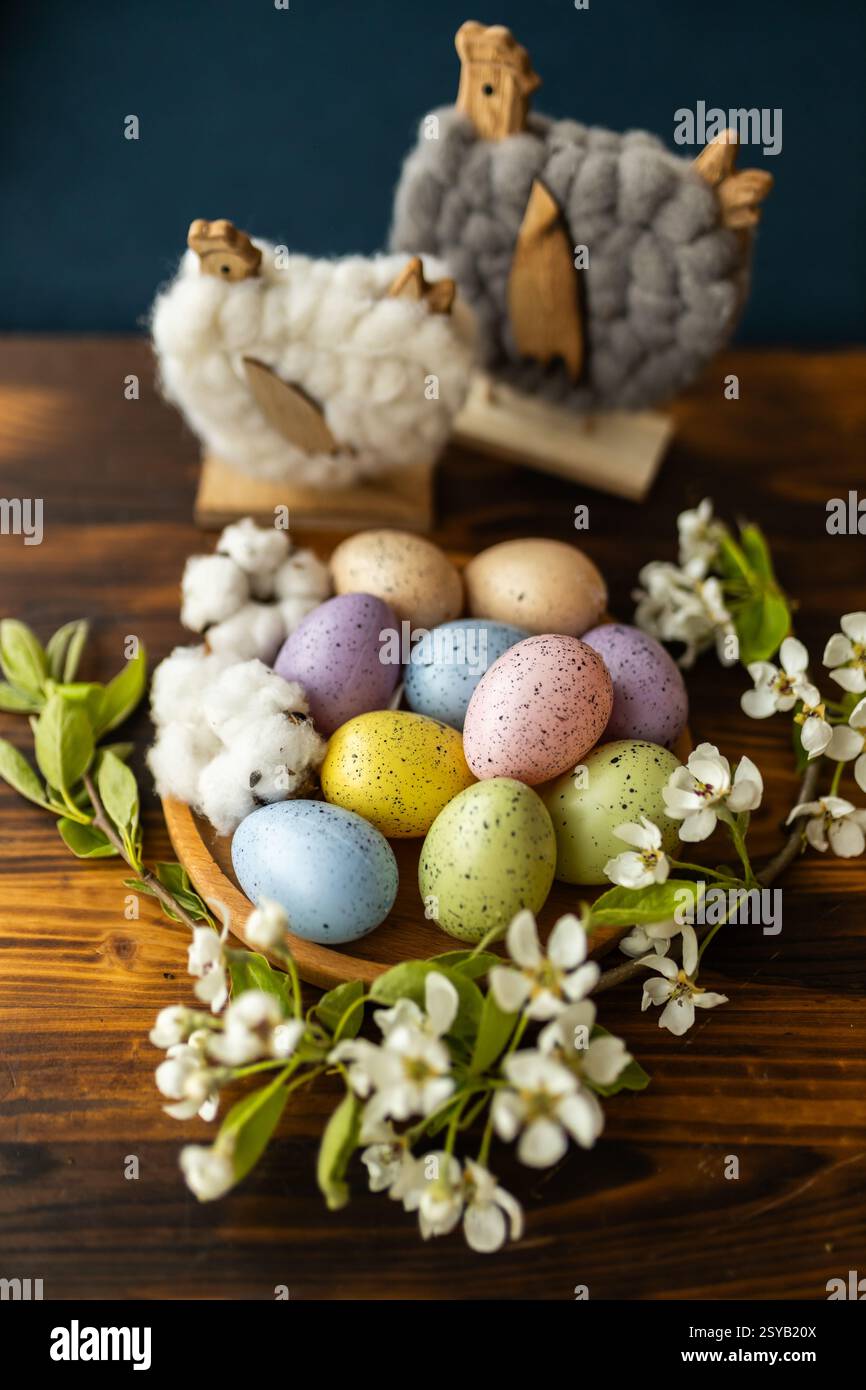 A rustic setting featuring colorful speckled Easter eggs in a wooden bowl, surrounded by white blossoms and woolly chickens decor on a wooden table. Stock Photo