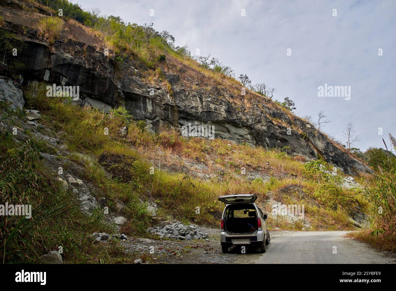 Photo of a car with its rear hatch open, parked on a dirt road beside a steep rocky cliff covered with dry grass and shrubs. Stock Photo
