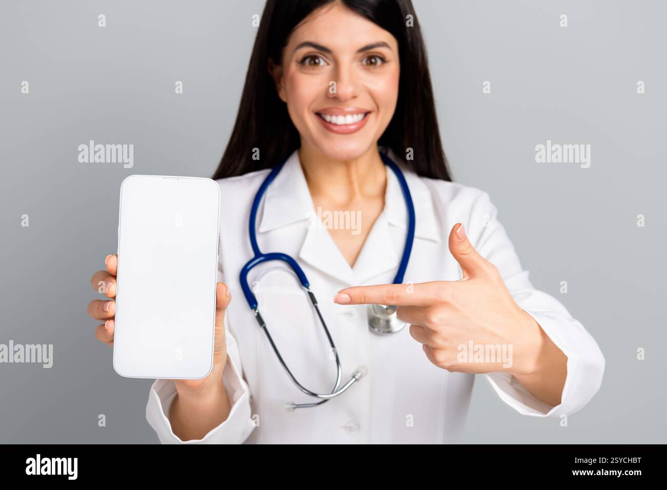 Smiling female doctor holding and pointing at smartphone with blank screen against grey background Stock Photo