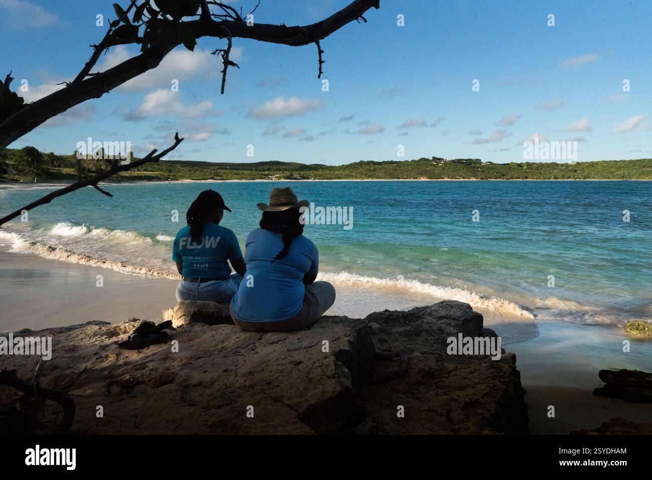 Two people sit on a rock in shade watching waves on shore at Half Moon Bay, Antigua Stock Photo