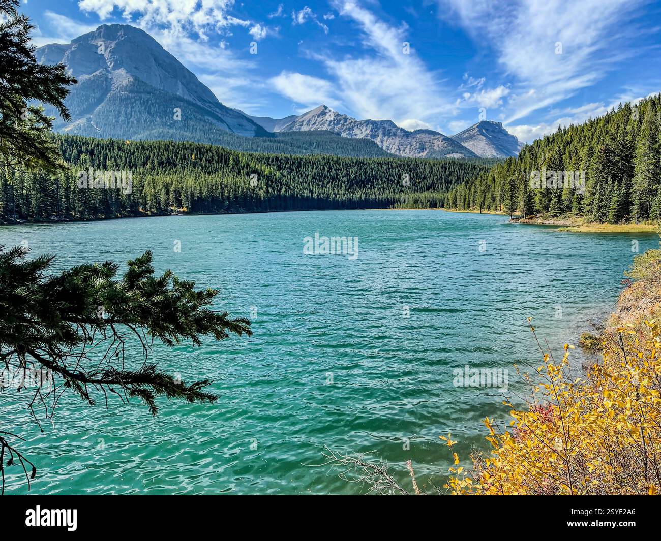 A turquoise lake sits at the base of a towering mountain range, surrounded by dense evergreen forests. The sky is bright blue with wispy clouds stretc - Smartphone Captured Stock Image
