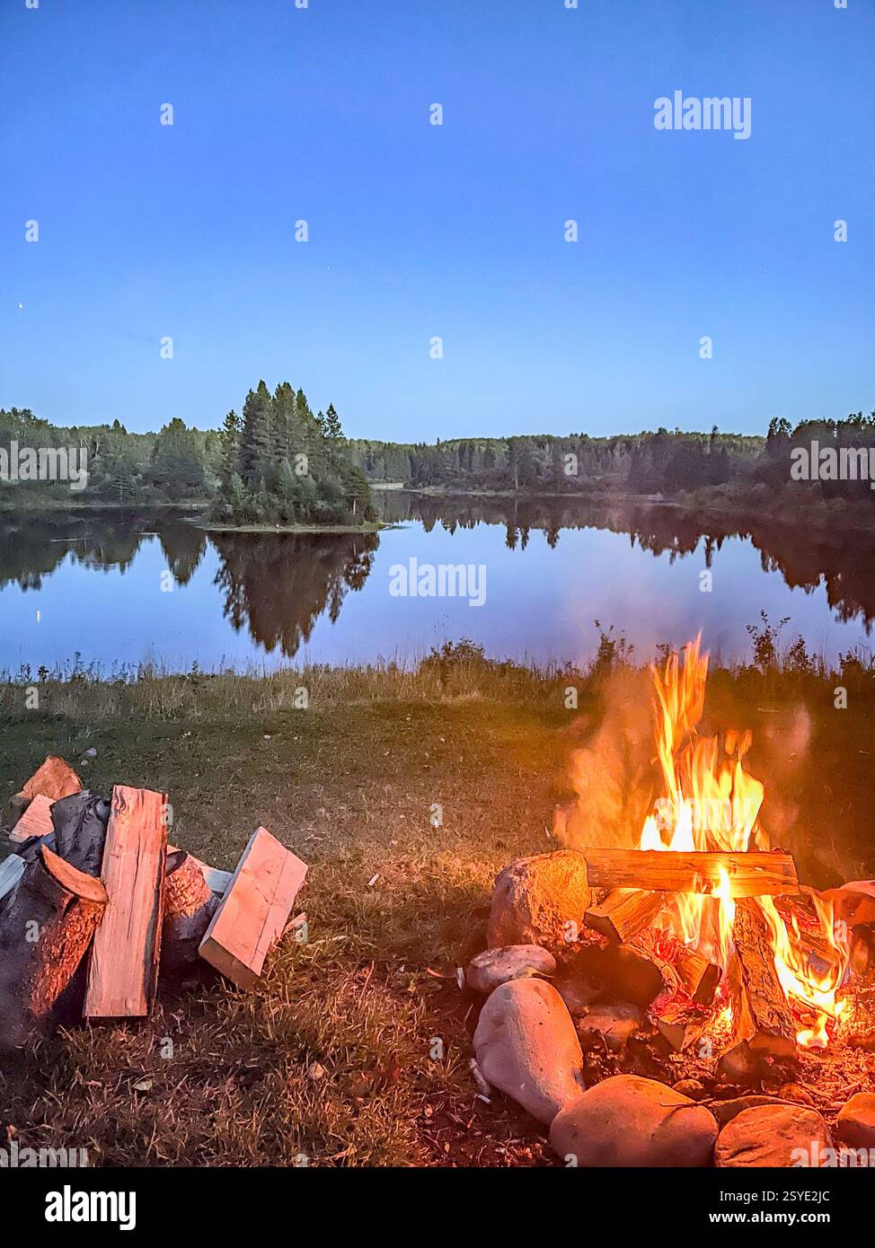 A warm campfire burns near a calm lake at dusk, with stacked firewood nearby. The flames contrast with the still water and soft twilight sky, creating - Smartphone Captured Stock Image