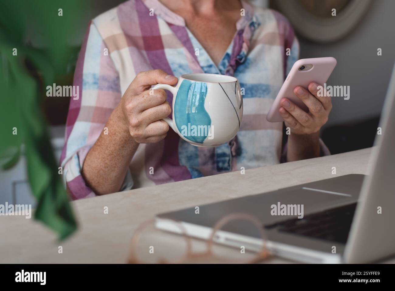 Woman sitting at desk in front of laptop computer holding cup of tea or coffee and using mobile phone Stock Photo