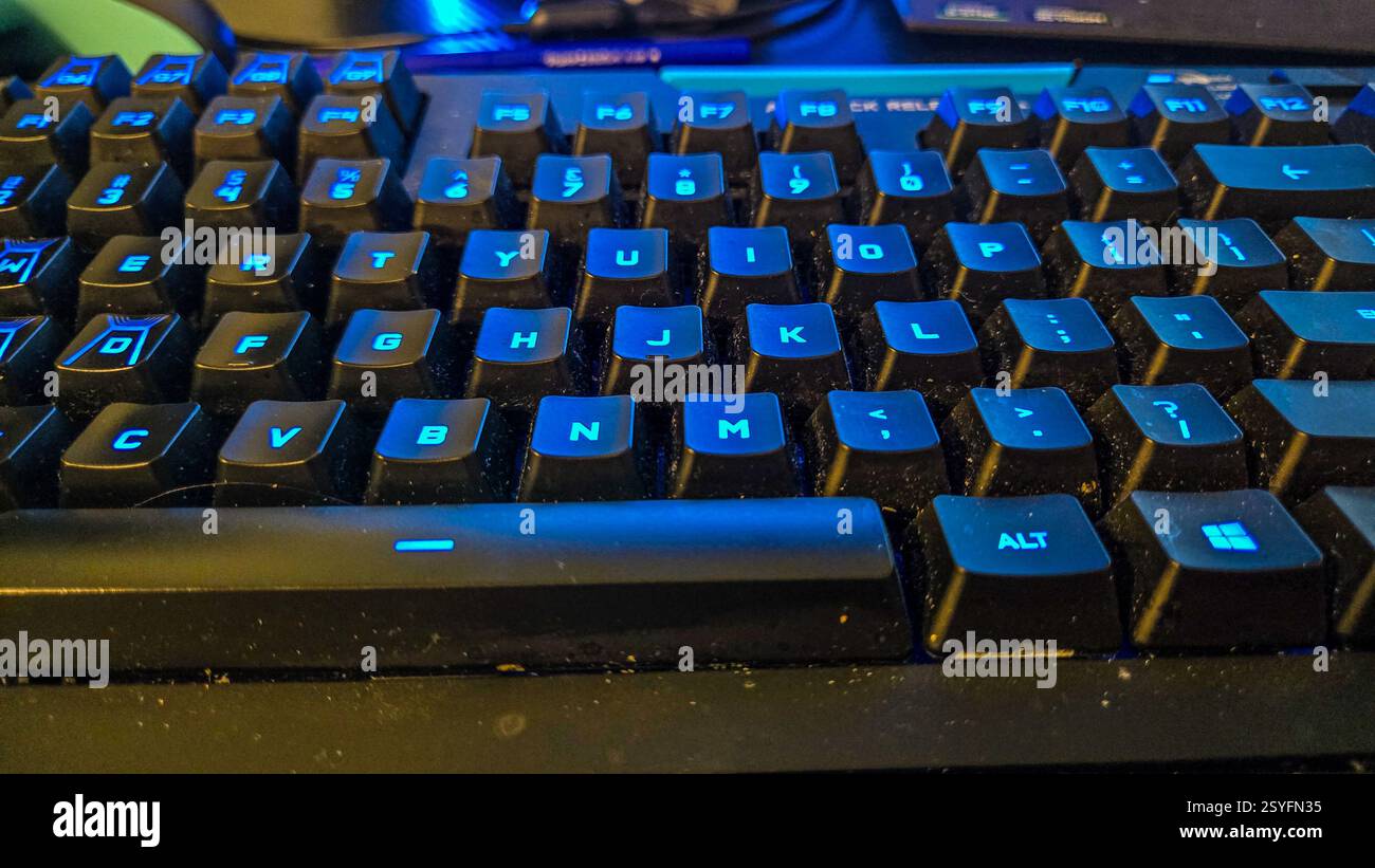 A close-up view of a keyboard displaying blue backlighting and noticeable dust on the keys. The dimly lit environment suggests late-night work or gami Stock Photo