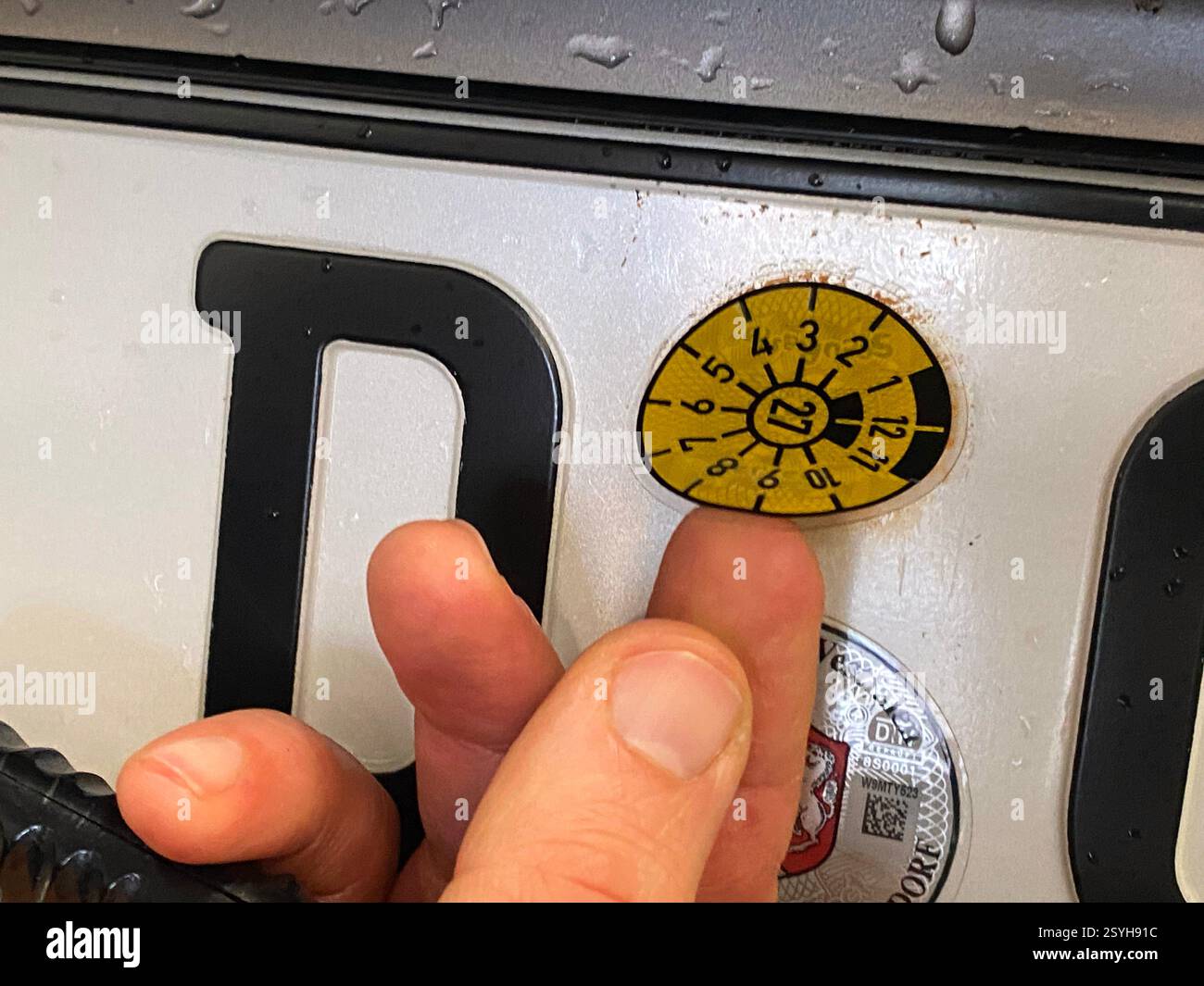 Eine frische TÜV-Plakette wird von dem Prüfer auf ein Autokennzeichen geklebt. Gültig ist die Plakette bis März 2027, die Farbe ist gelb. - Smartphone Captured Stock Image