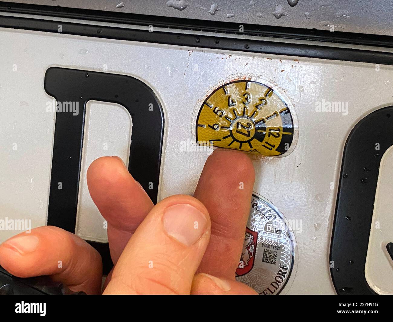 Eine frische TÜV-Plakette wird von dem Prüfer auf ein Autokennzeichen geklebt. Gültig ist die Plakette bis März 2027, die Farbe ist gelb. - Smartphone Captured Stock Image