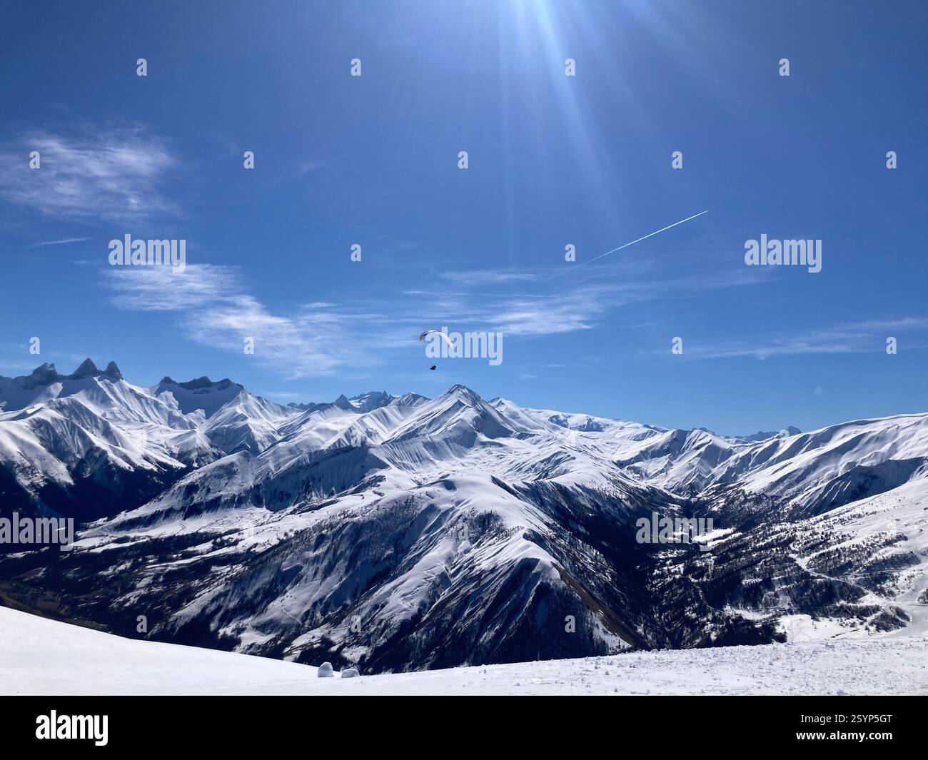 A paraglider soars over a snowy Alpine valley bathed in golden light, with majestic peaks in the background under a clear blue sky. - Smartphone Captured Stock Image