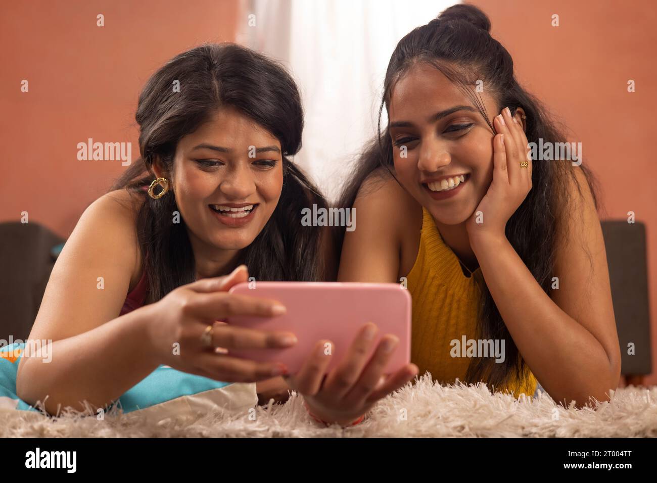 Close-up portrait of two smiling women together watching video on mobile phone while lying on floor in living room Stock Photo