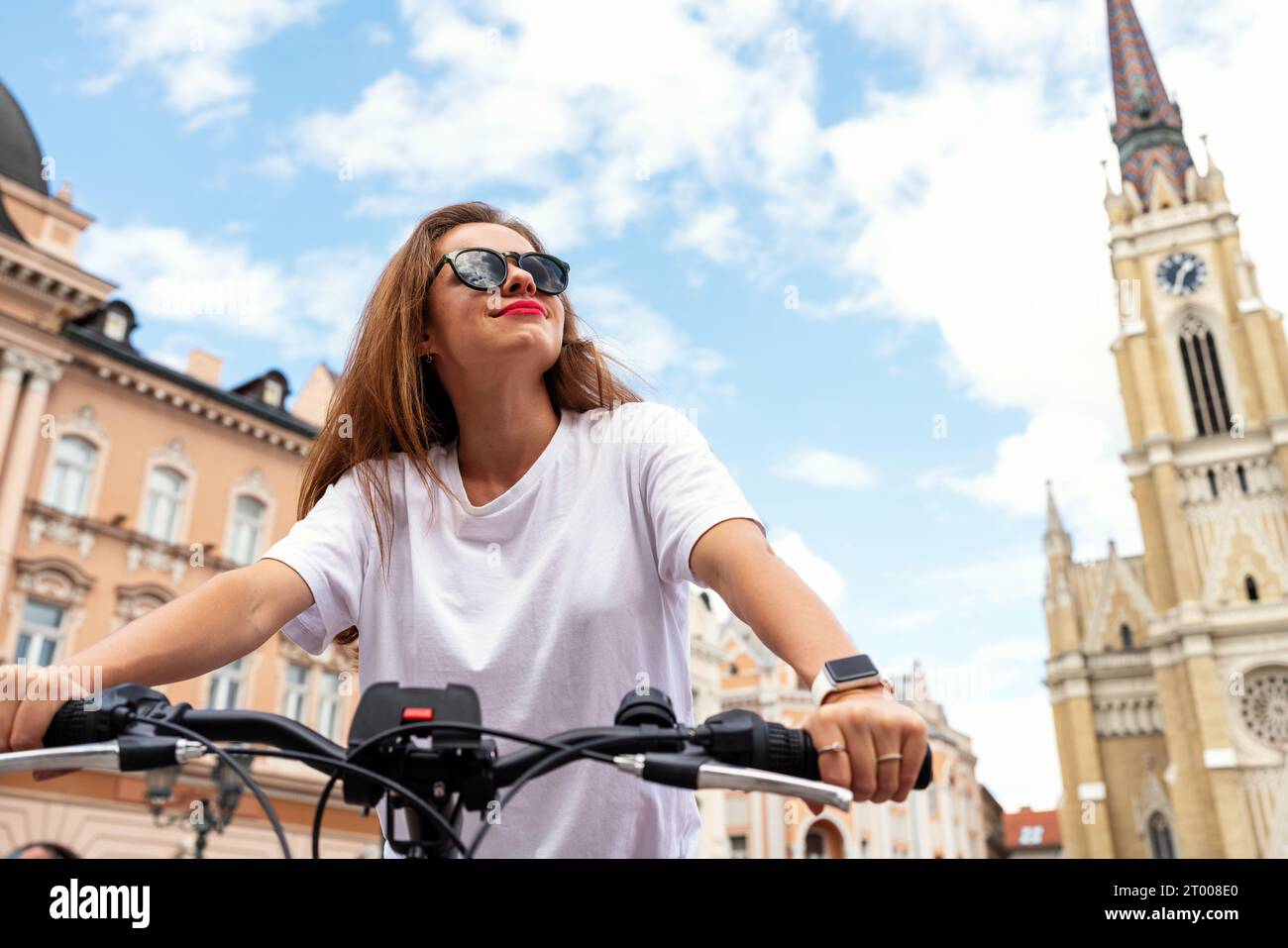 Brunette woman wearing sunglasses riding a bike, bicycle travel tour around historic place in Novi Sad, Serbia. Stock Photo