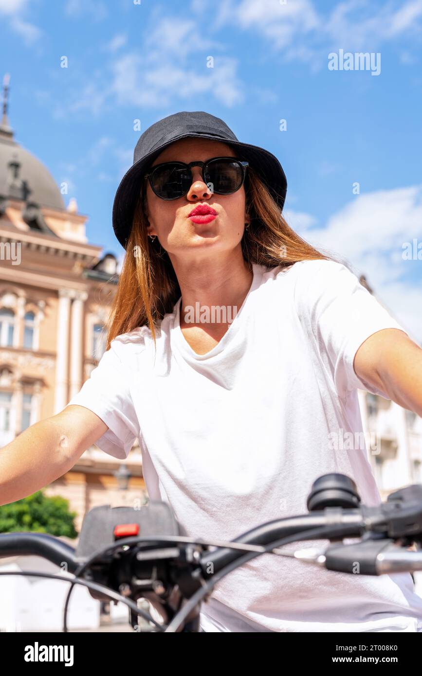 Stylish young woman wearing hat and sunglasses enjoys cycling around the city in summer. Red lipstick lips Air kissing. Stock Photo