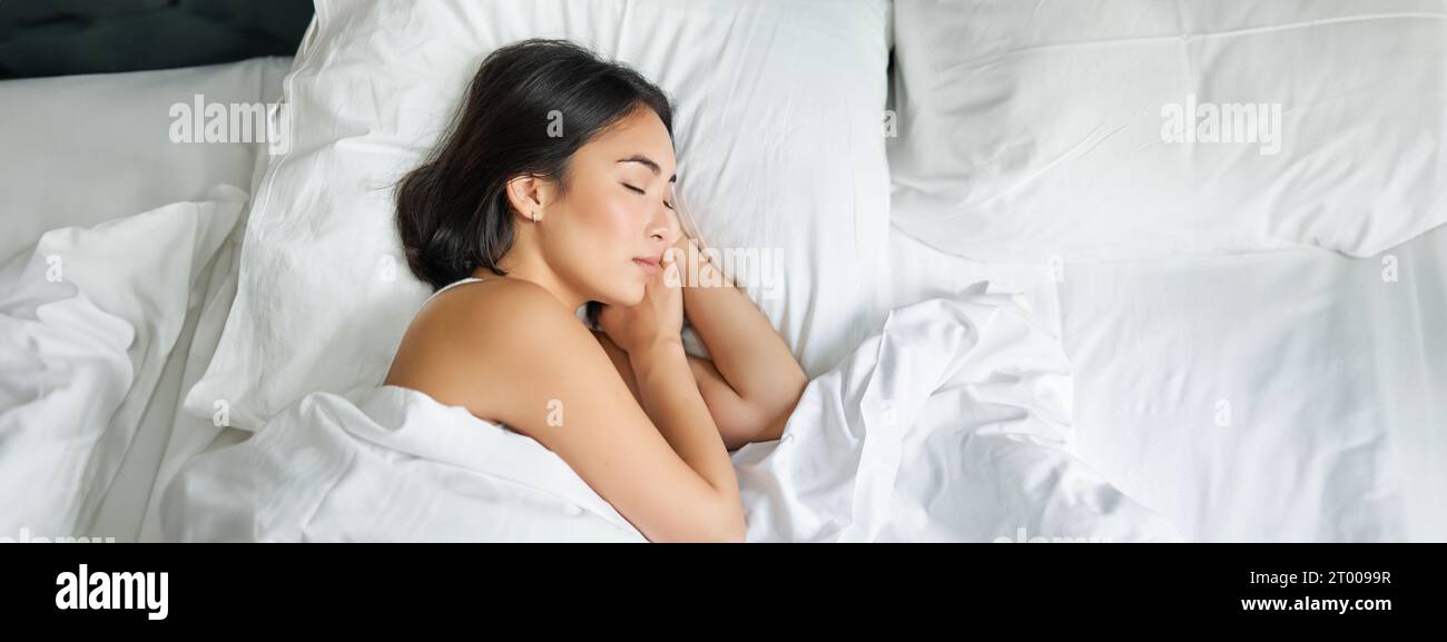 Top view image of asian woman sleeping alone in king size bed on white pillows. Young girl lying in her bedroom with eyes closed, morning sunlight Stock Photo