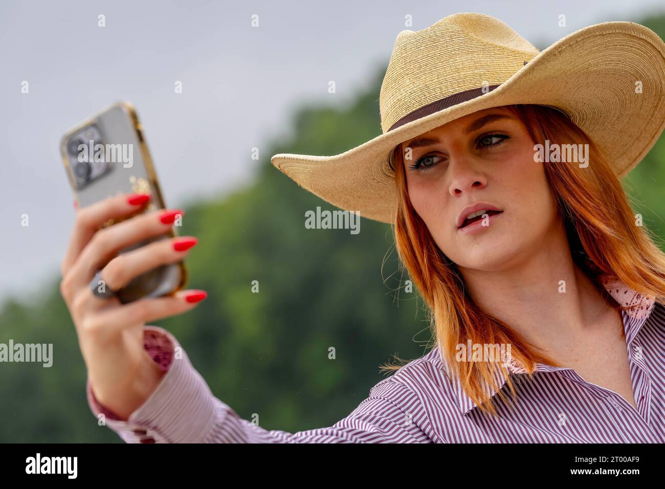A Lovely Red Headed Cowgirl Uses Her Cellphone In A Country Western Setting Outdoors Stock Photo