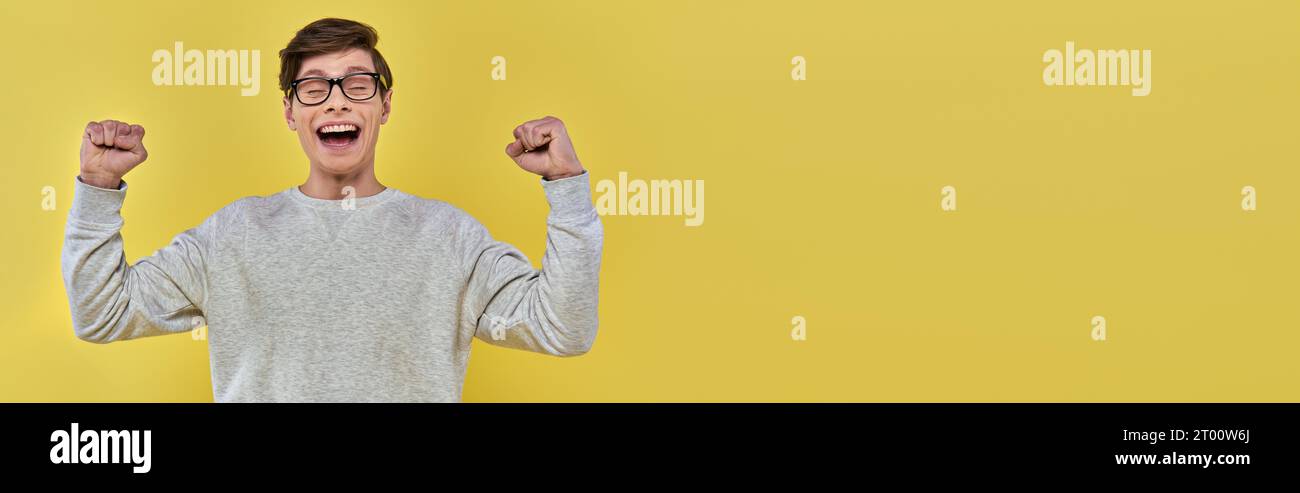 cheerful young man  in white sweatshirt and glasses raising fists up and closing eyes, banner Stock Photo