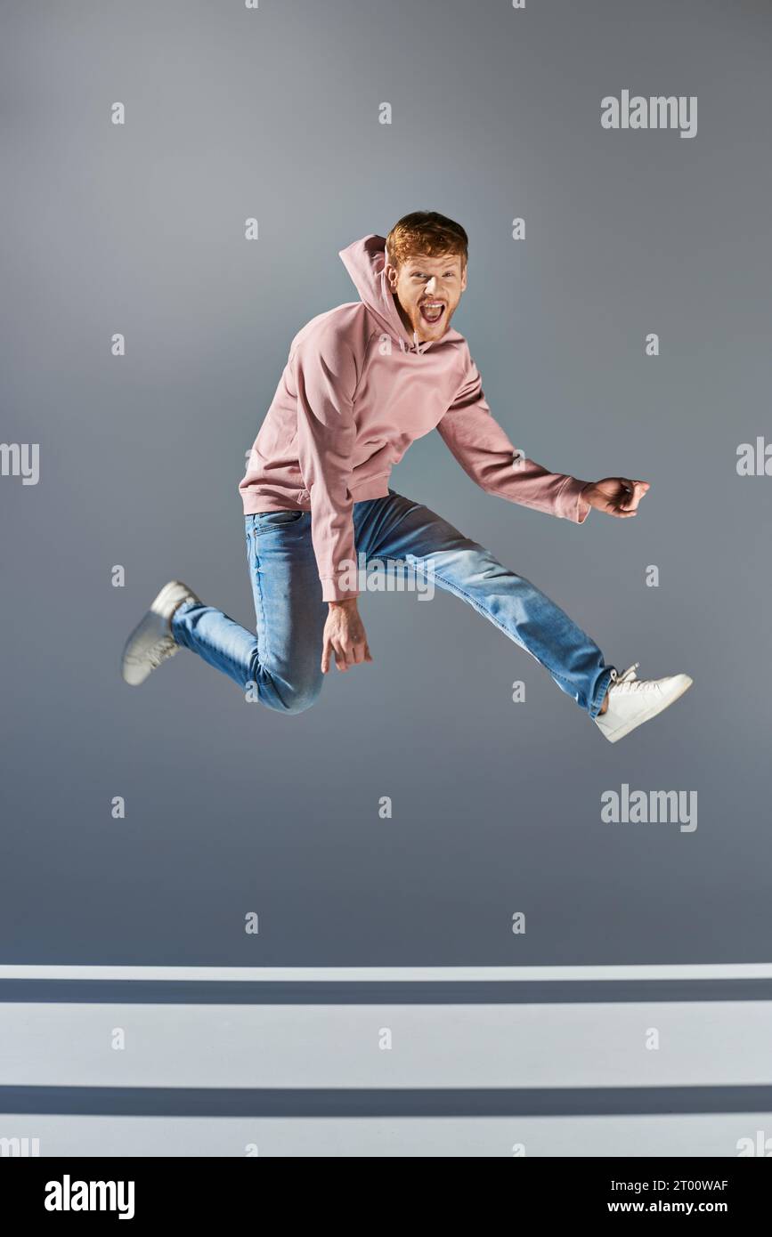cheerful young man in casual clothing jumping and playing invisible guitar on grey backdrop Stock Photo