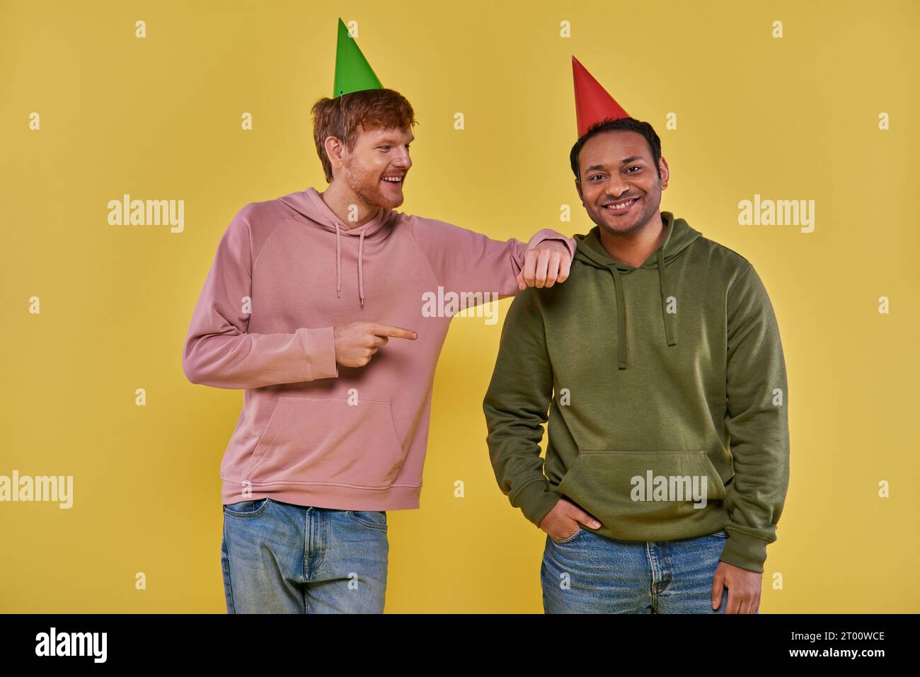 two cheerful men in casual attire and birthday hats posing together on yellow backdrop, birthday