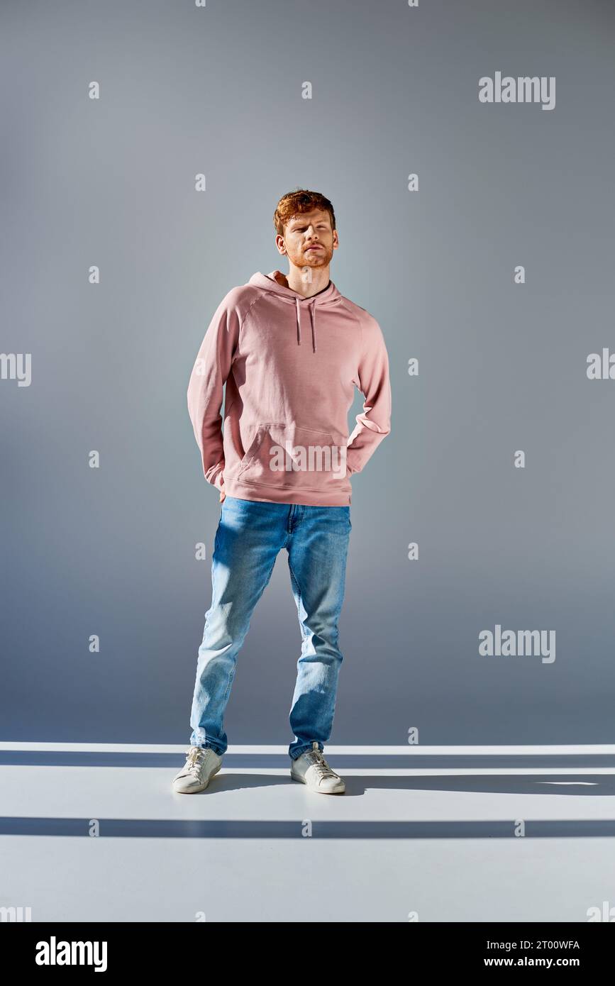 handsome red haired man in pink hoodie posing with hands in pockets of jeans on grey backdrop Stock Photo