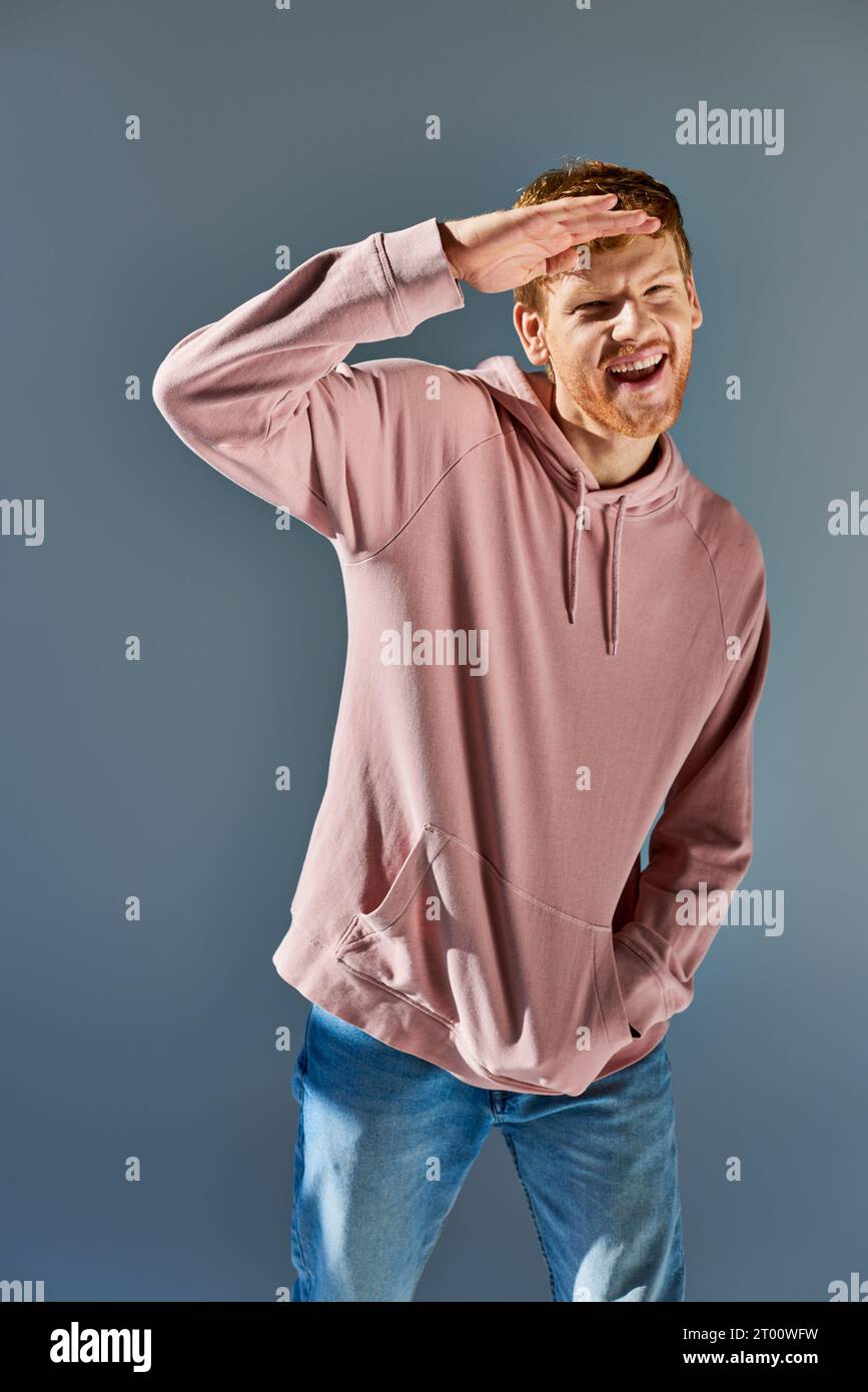 cheerful young man in trendy hoodie and jeans holding hand at forehead posing on grey backdrop Stock Photo