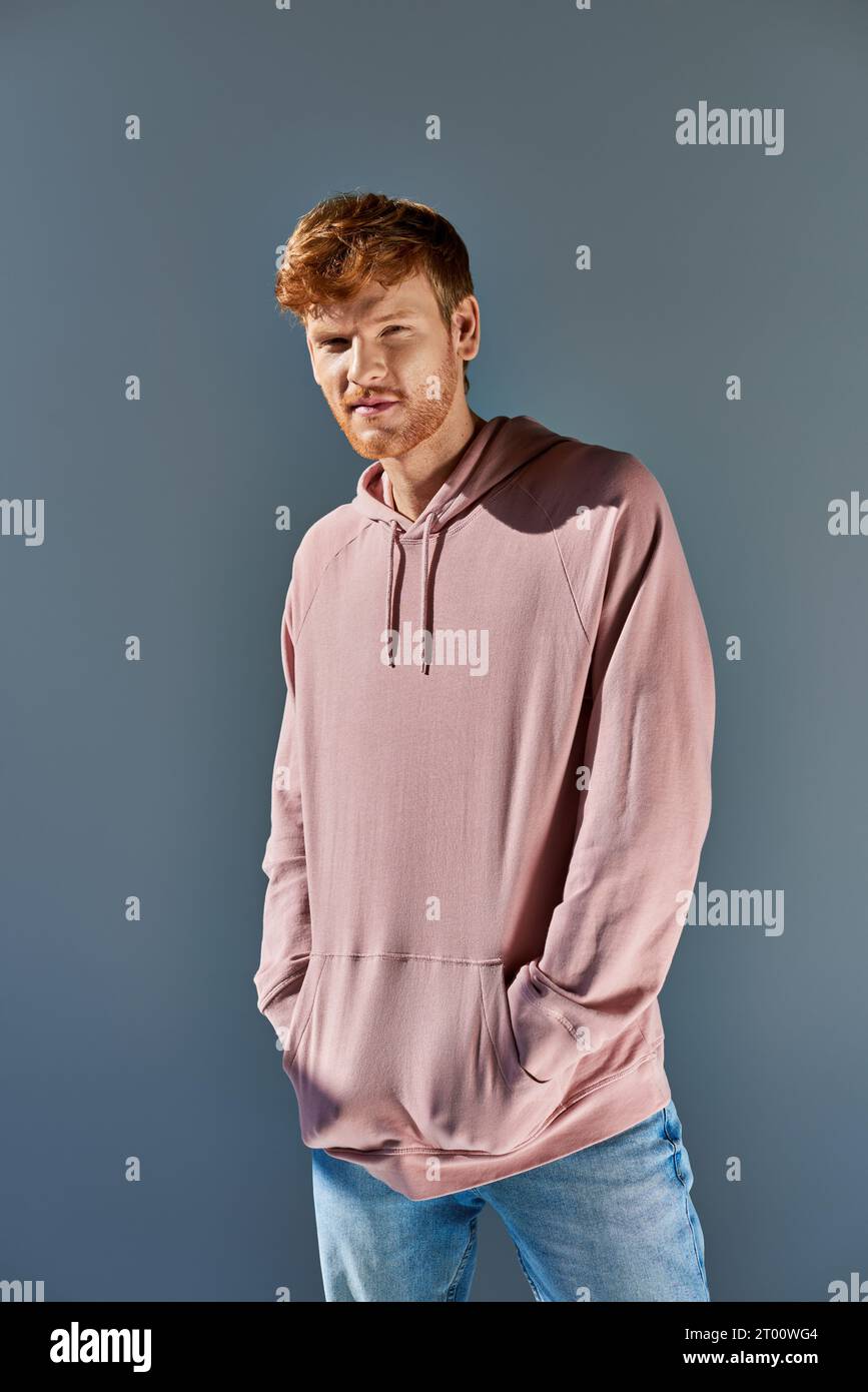 stylish young red haired man in pink hoodie and jeans looking at camera posing on grey backdrop Stock Photo