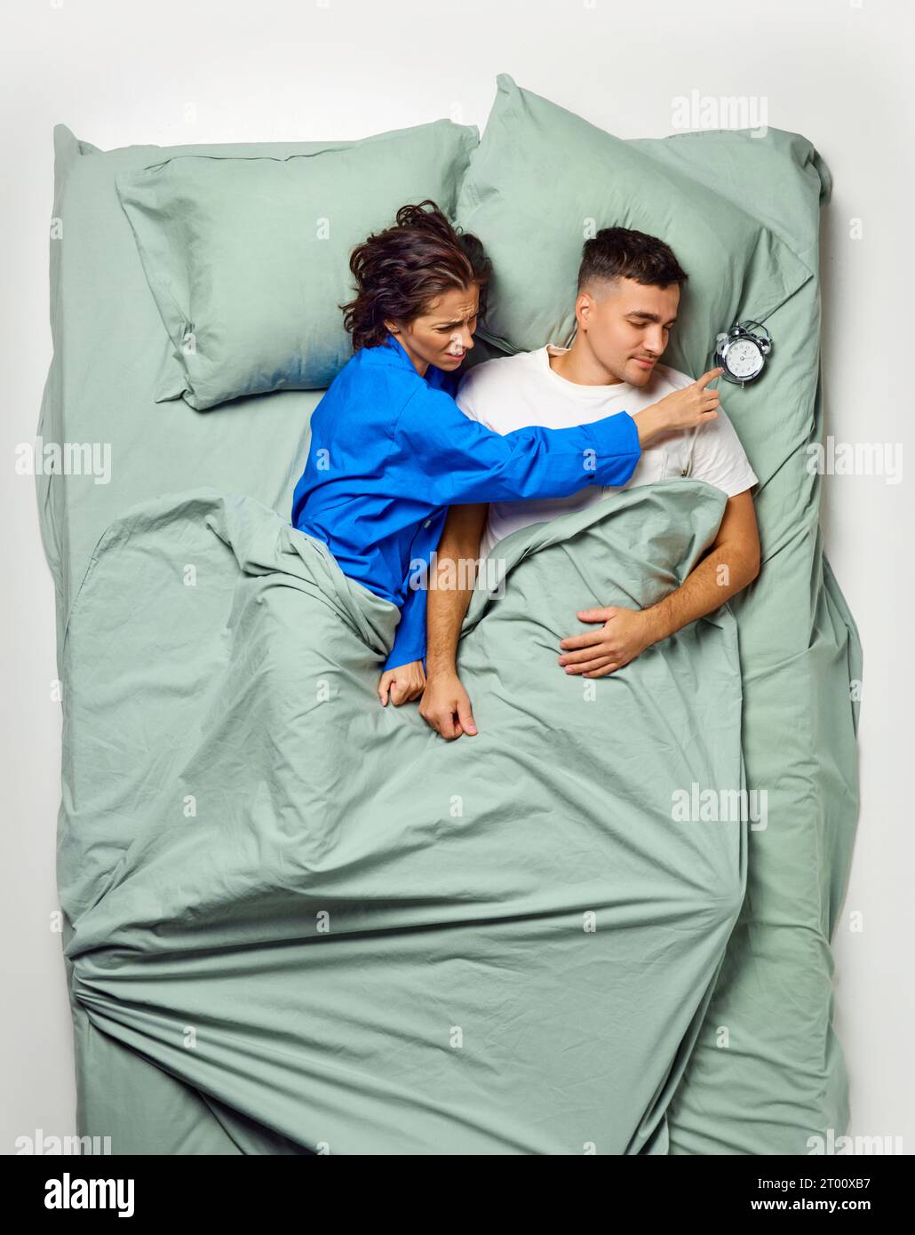 Young beautiful woman reaching across attractive man lying on back for turning of loud alarm clock, couple relaxing in big mint bed. Overslept. Stock Photo