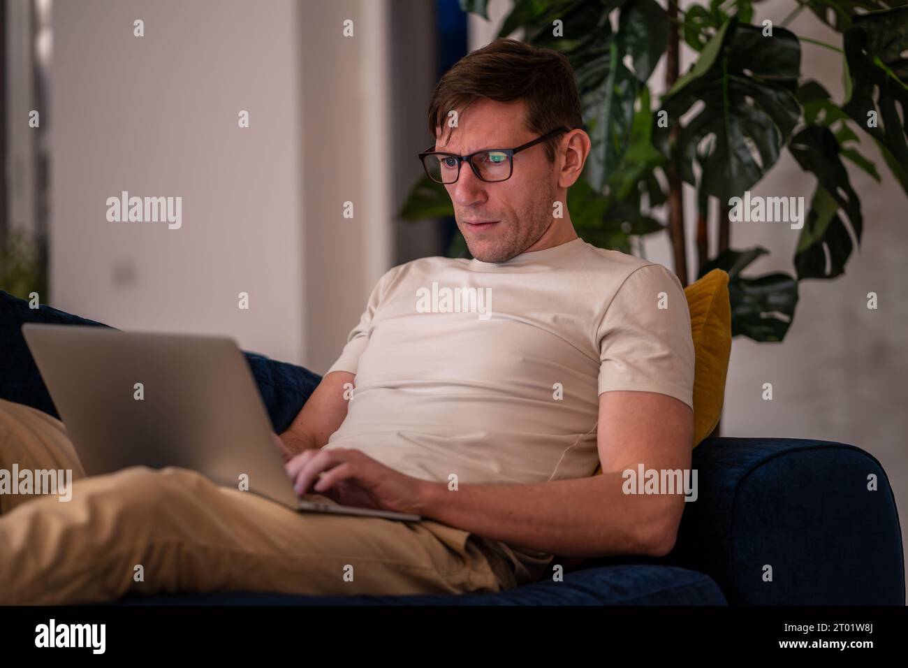 Man middle aged student working on educational project lying on couch at home in evening after job. Stock Photo