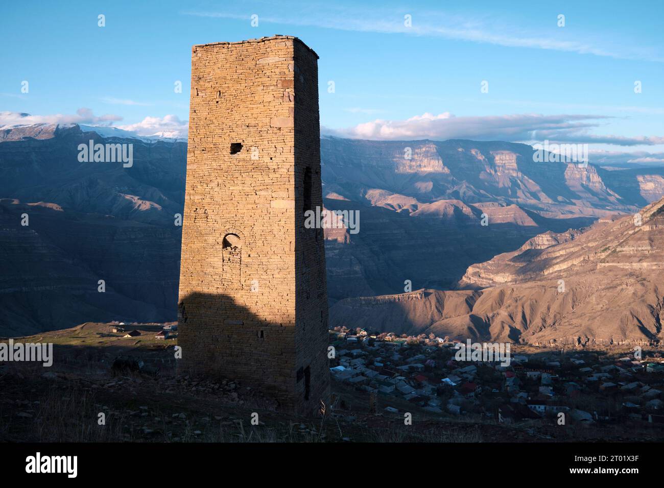 The ancient town tower in the village stands on the vibrant sunset. In the Dagestan Republic, during the beautiful golden hour, a charming old town tower in the village of Goor stands against the backdrop of stunning mountains, all bathed in the warm colors of the sunset. Stock Photo