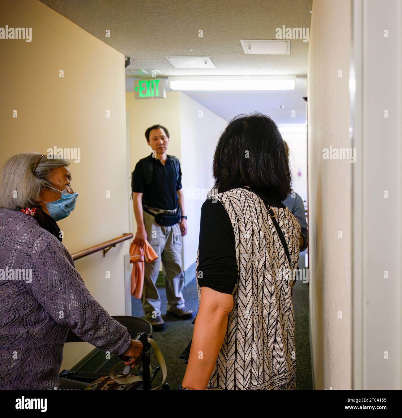 Senior woman with facemask walking using a mobility walker in the apartment corridor, accompanied by her son and daughter. Stock Photo