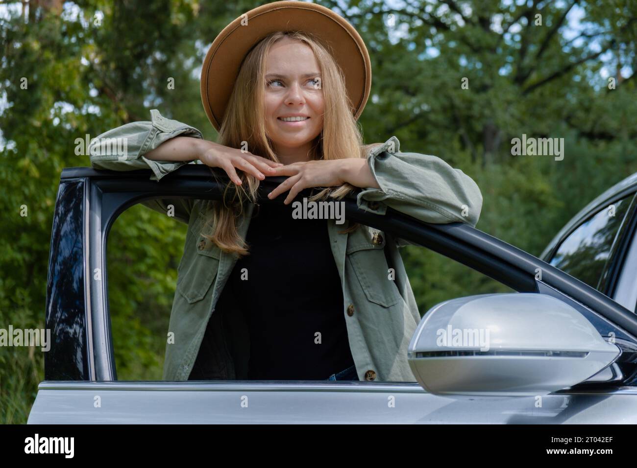 Blonde woman in hat staying next to car door. Young tourist explore local travel making candid real moments. True emotions expressions of getting away and refresh relax on open clean air Stock Photo