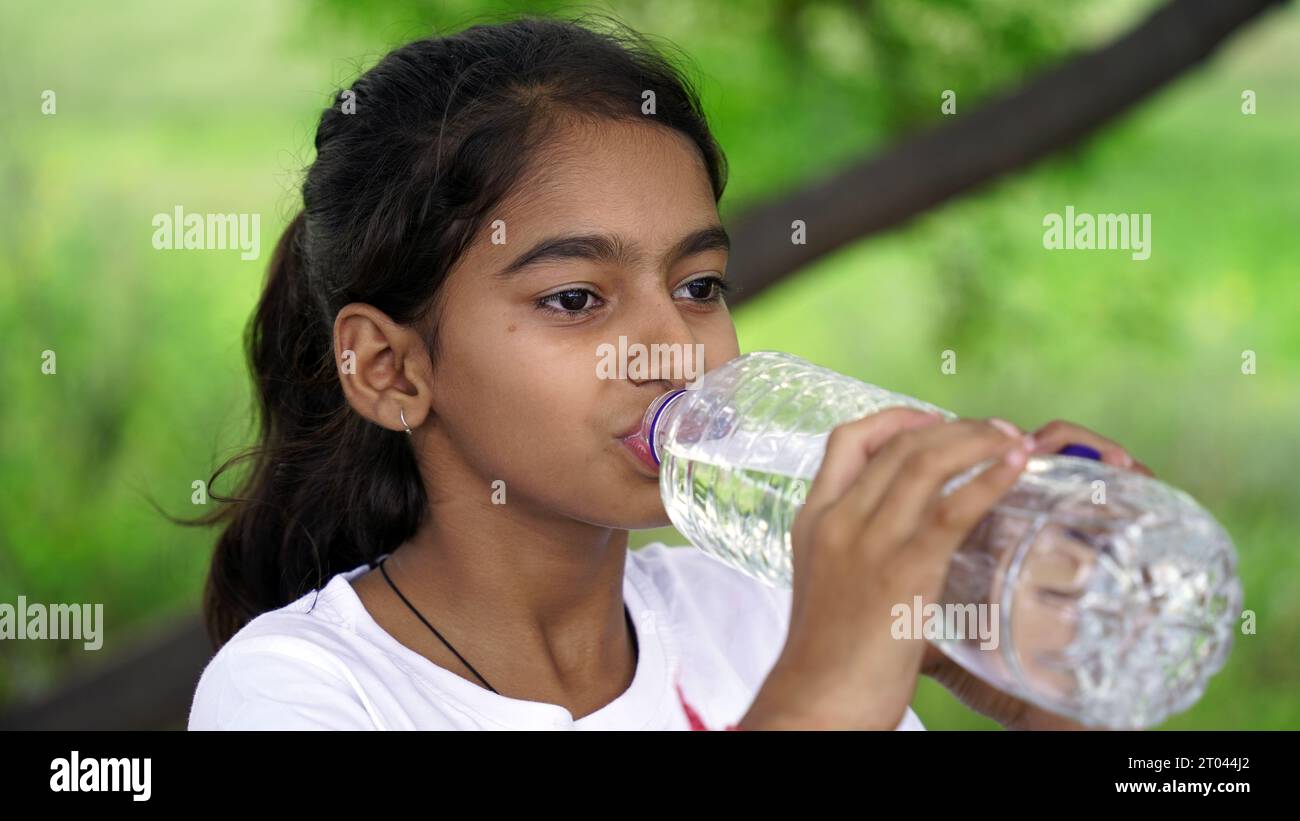 Happy Cute Indian girl with black hair drinking water in the park, holding plastic bottle and showing thumb up. outdoor shot Stock Photo