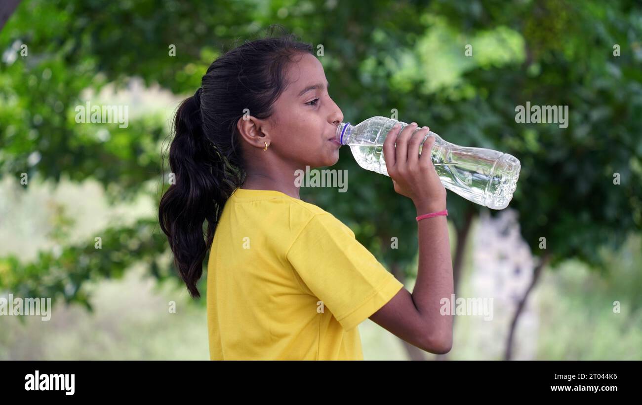 Happy Cute Indian girl with black hair drinking water in the park, holding plastic bottle and showing thumb up. outdoor shot Stock Photo