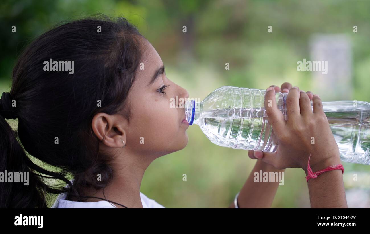 Happy Cute Indian girl with black hair drinking water in the park, holding plastic bottle and showing thumb up. outdoor shot Stock Photo