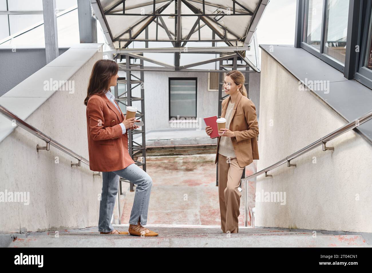 two colleagues in business casual attire standing on stairs smiling at each other, coworking concept Stock Photo