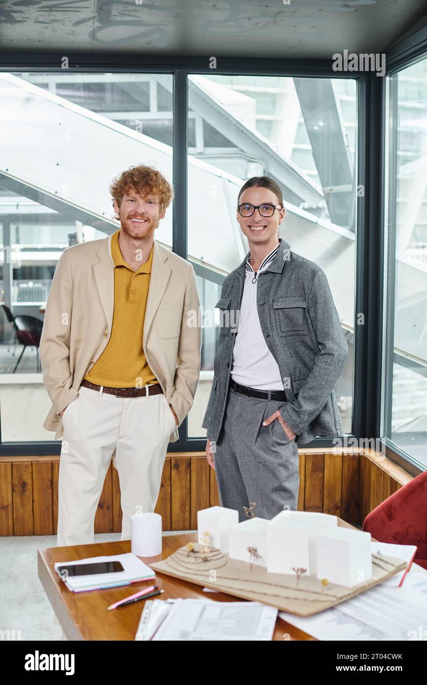 two male coworkers posing near table with scale model and looking at camera, design bureau Stock Photo