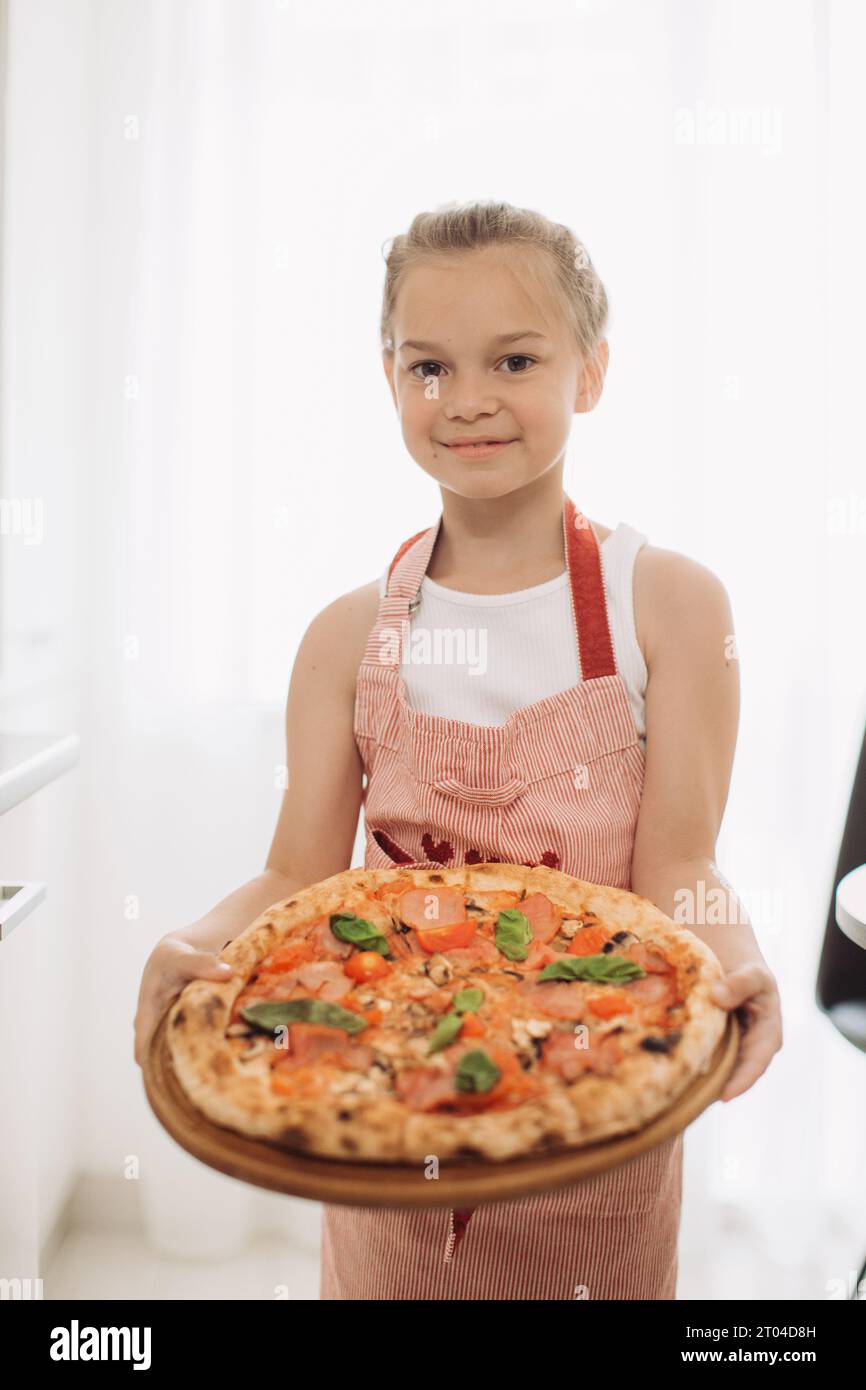 A little 8-year-old girl cook holds a delicious pizza while standing in