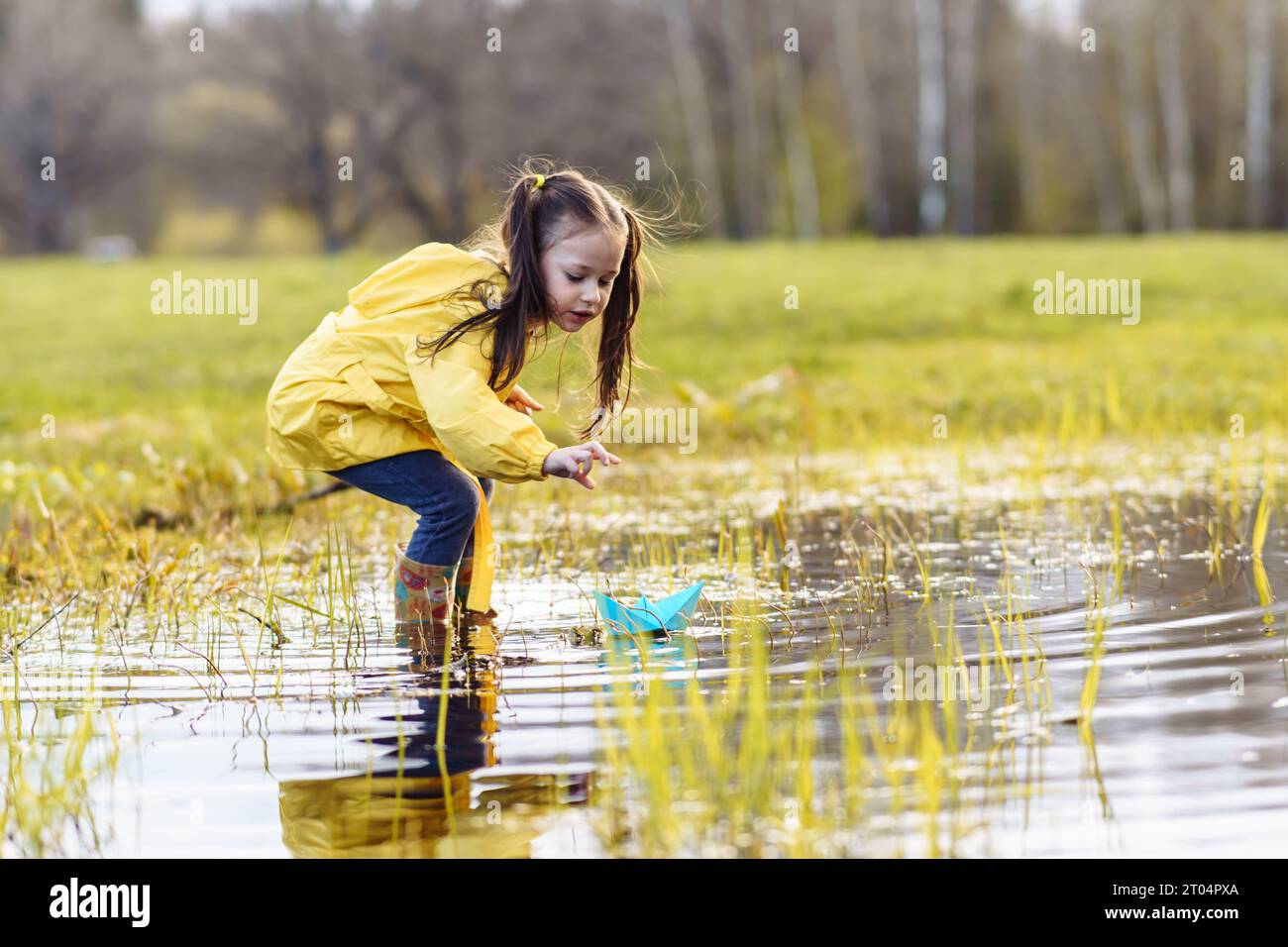 Charming little girl in yellow jacket and rubber boots standing in puddle on green lawn, stretching arm forward and bending over, carefully looking at Stock Photo