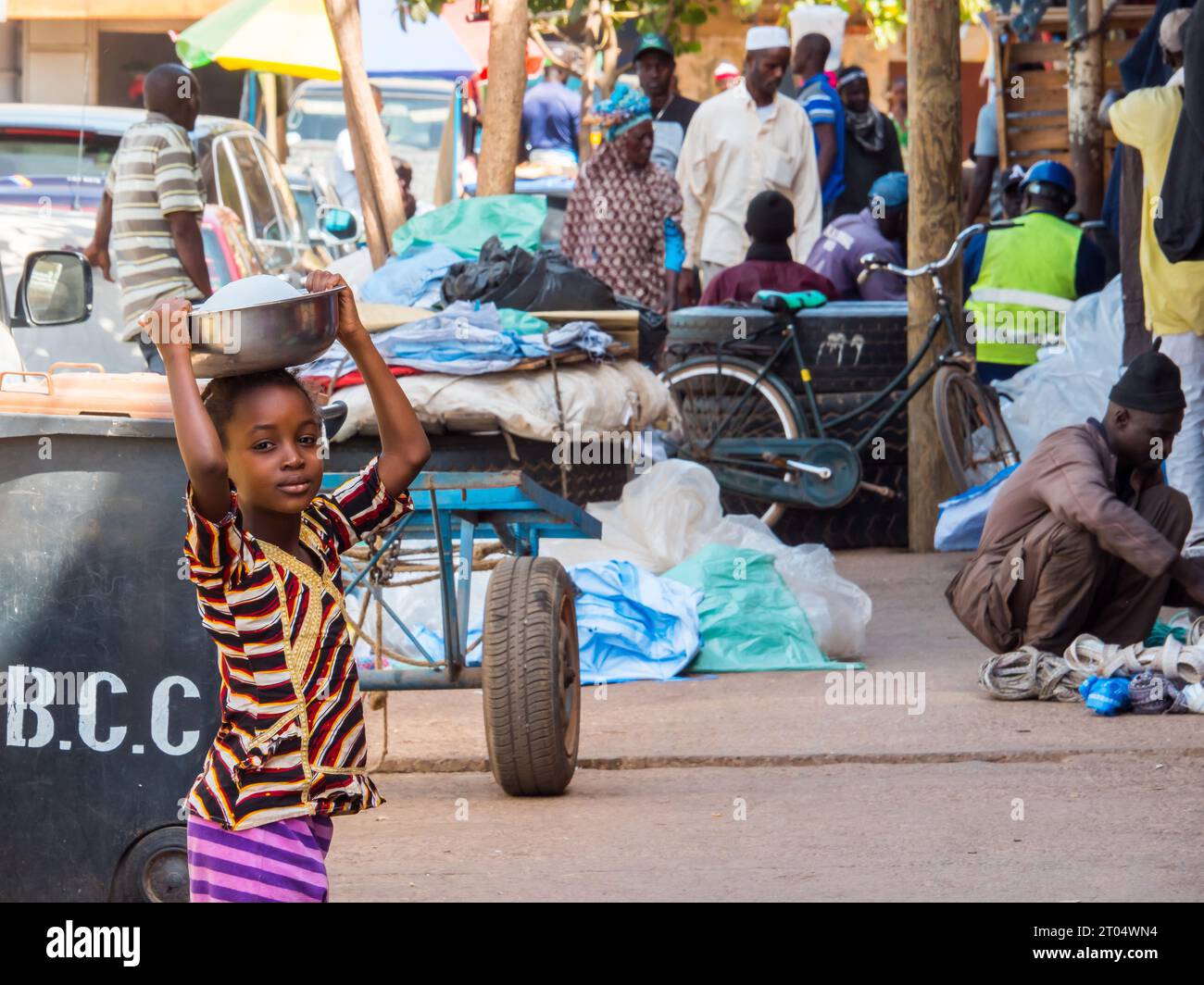 child with a bowl on the head on a local market in a small town., Gambia Stock Photo