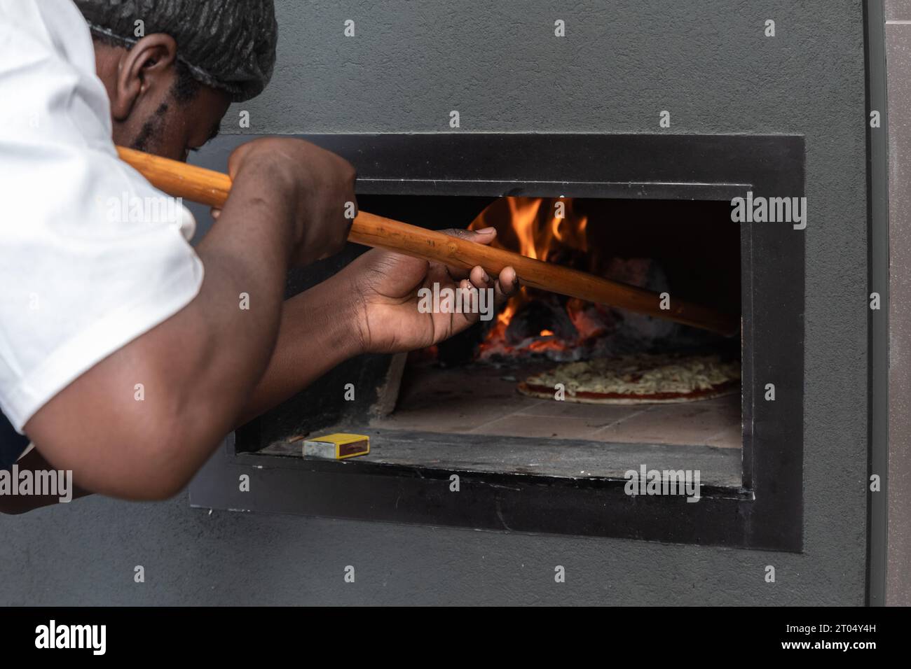 In the intense glow of the pizza oven's flames, a skilled black man, using a wooden peel, expertly maneuvers a sizzling pizza Stock Photo