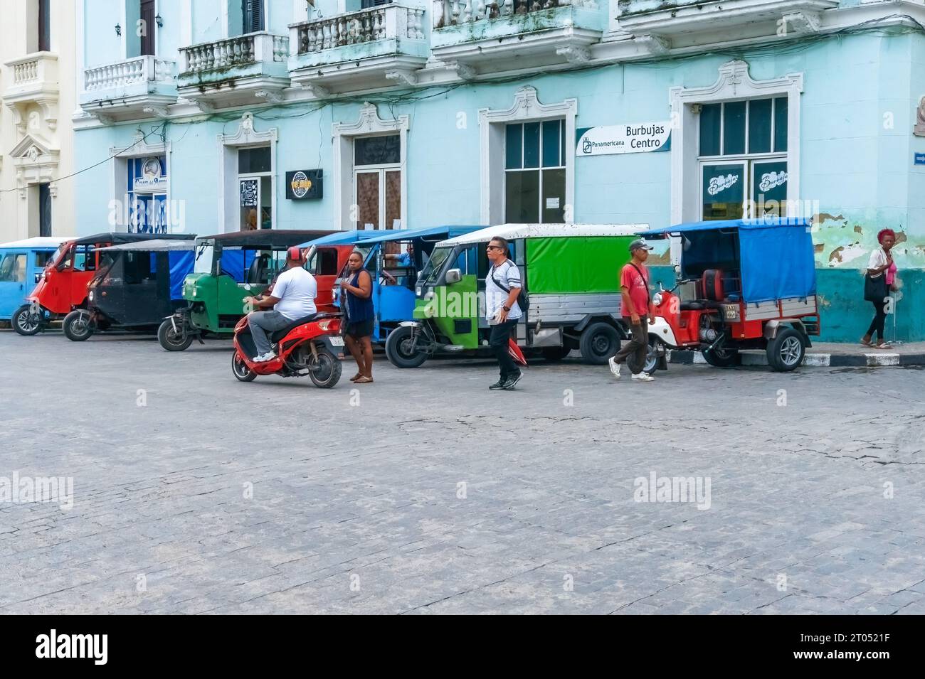 A Cuban man and other people are by vintage tricycles or motonetas. The motor vehicles are used in local urban transportation. They are privately oper Stock Photo