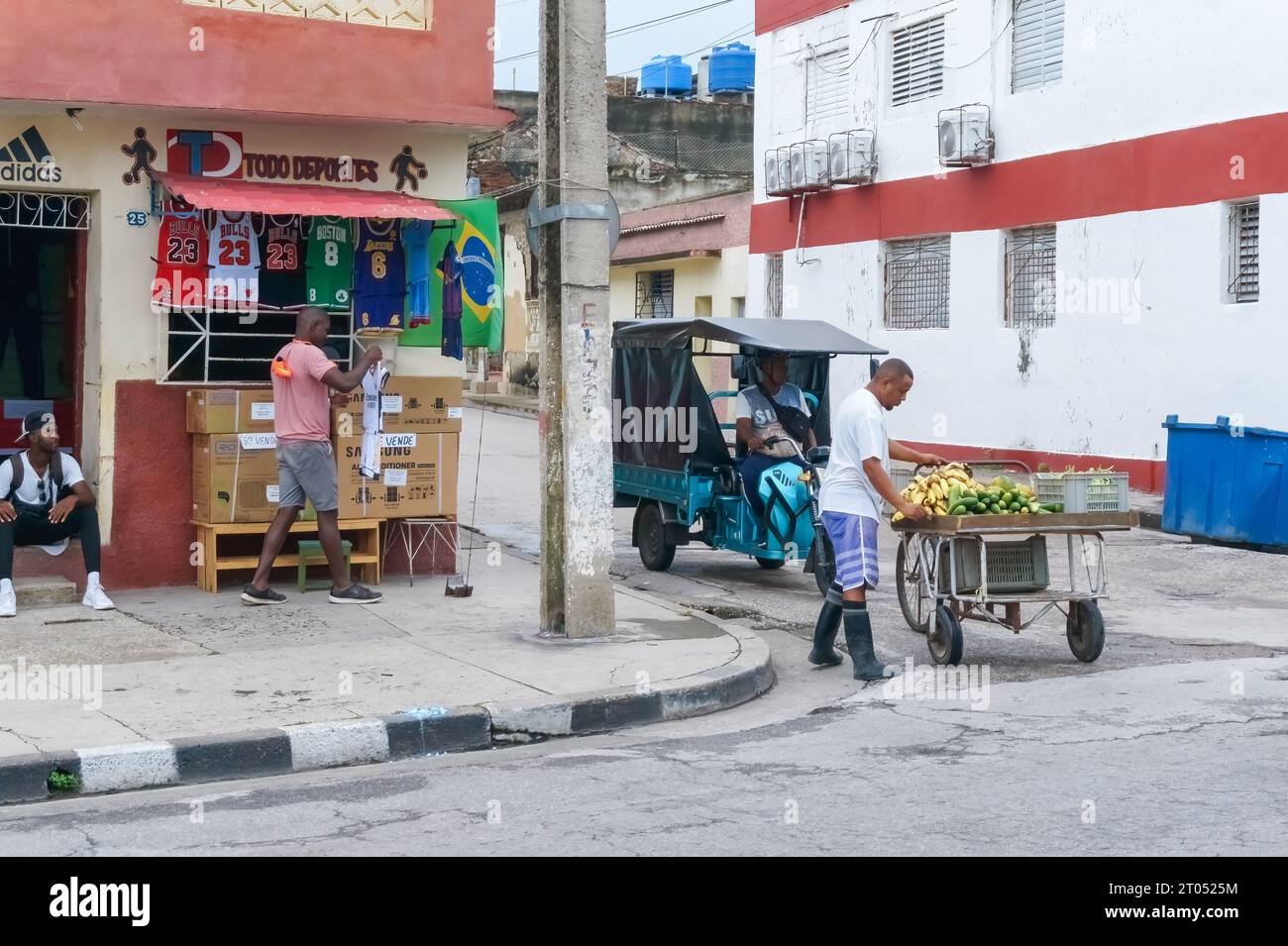 A Cuban man sells fruits and vegetables in a cart at a street corner. Another male person sets his house kiosk with sports items for sale. Santa Clara, Stock Photo